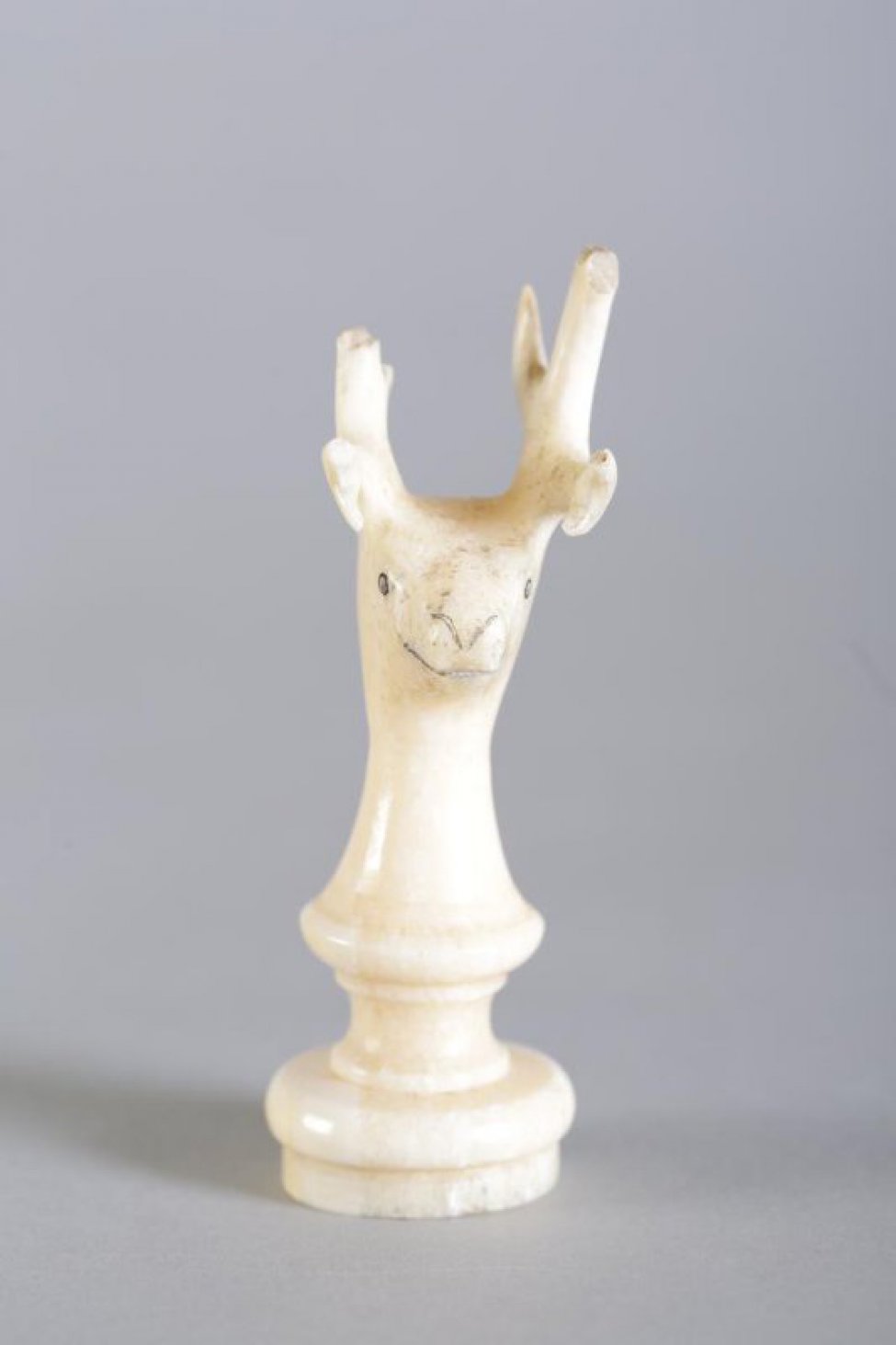 Шахматная фигура белого цвета в виде невысокой балясины, вверху заканчивающаяся головой рогатого оленя. Маленькие круглые глаза, линия рта и кончик носа обозначены неглубокой резьбой (процарапыванием) с черной тонировкой. Вся фигура выполнена из одного куска бивня.