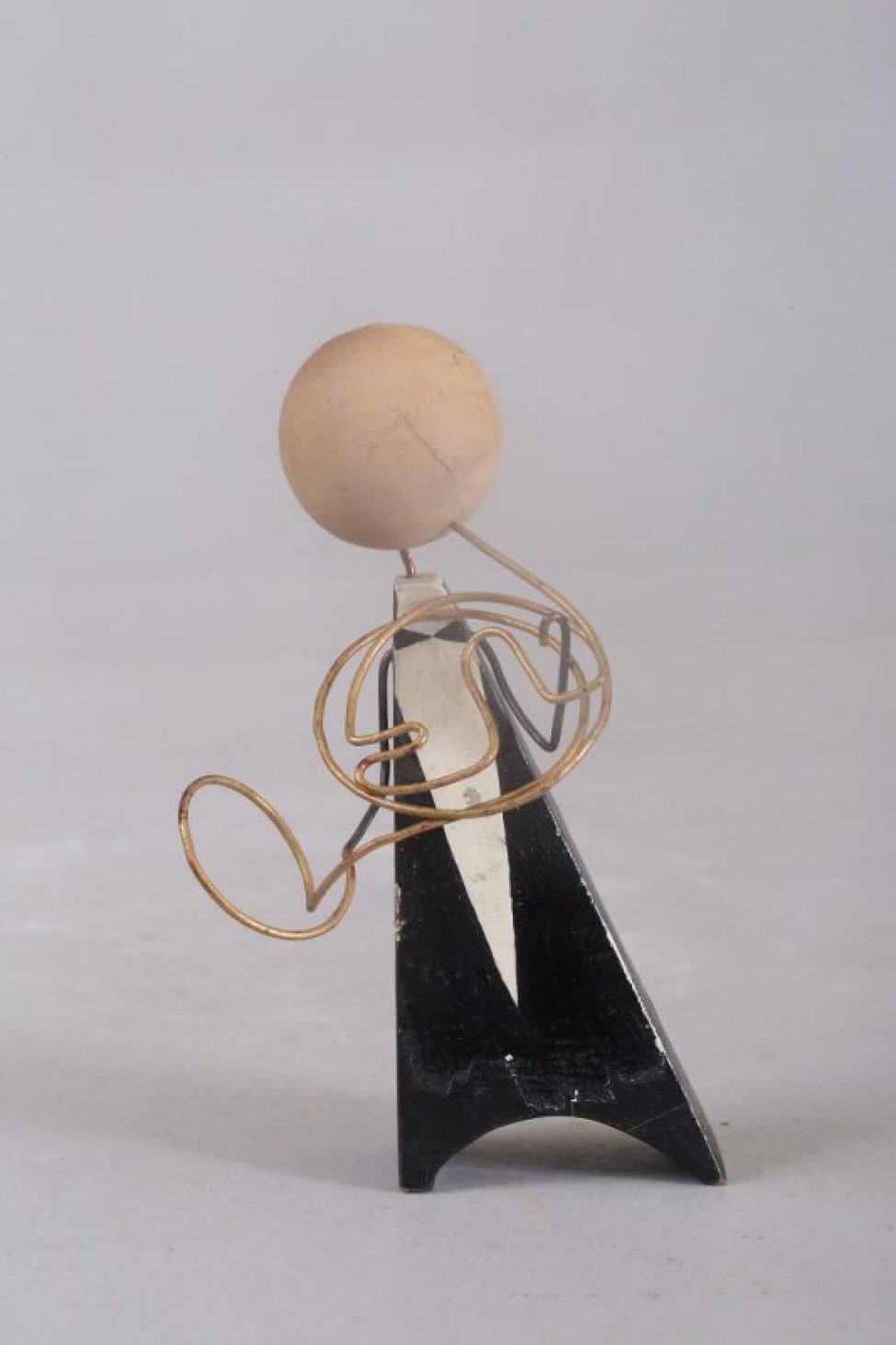 Фигурка в виде черного треугольника с полукруглой выемкой внизу, с белой клинообразной полоской, с головой в виде желтого шарика. Инструмент и руки из проволоки.